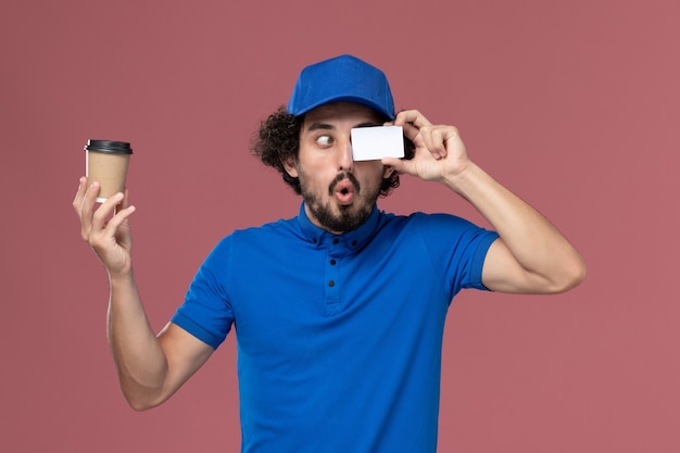 Front view of male courier in blue uniform and cap with delivery coffee cup and card on his hands on the pink wall