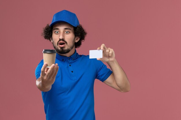 青い制服とピンクの壁に彼の手に配達コーヒーカップとカードとキャップの男性宅配便の正面図