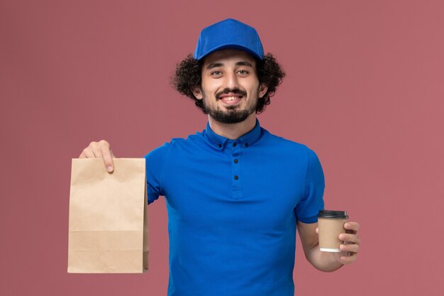 Front view of male courier in blue uniform and cap with delivery coffee cup and food package on his hands on pink wall