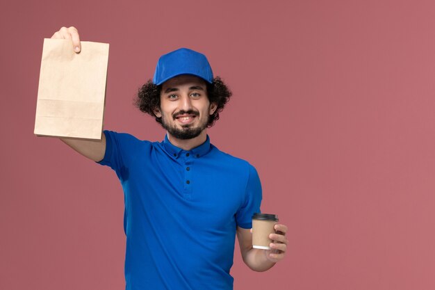 Вид спереди курьера-мужчины в синей форме и кепке с доставкой кофейной чашки и продуктового пакета на руках на розовой стене