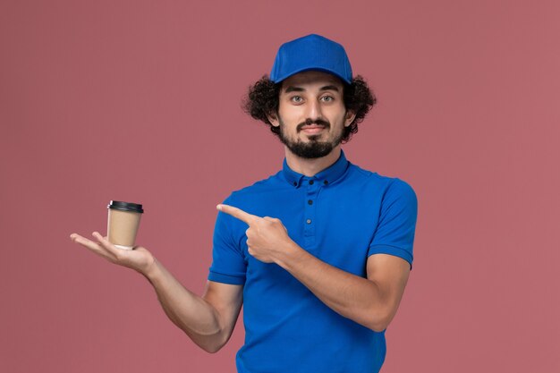 Front view of male courier in blue uniform and cap with delivery coffee cup on his hands on pink wall