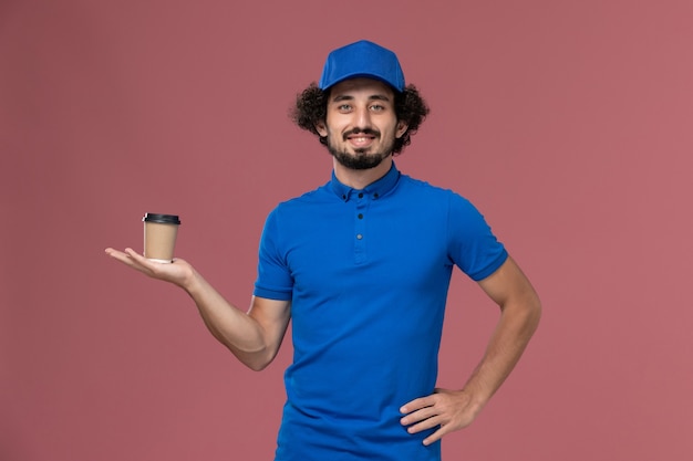 Front view of male courier in blue uniform and cap with delivery coffee cup on his hands on pink wall