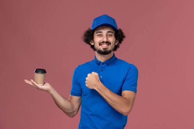 Вид спереди курьера-мужчины в синей форме и кепке с доставкой кофейной чашки на руках на розовой стене