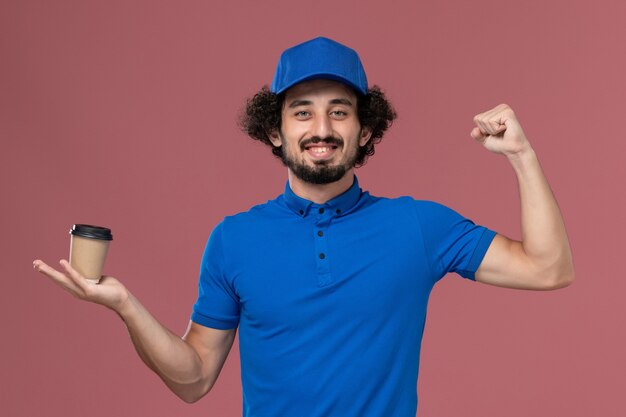 Front view of male courier in blue uniform and cap with delivery coffee cup on his hands on pink wall