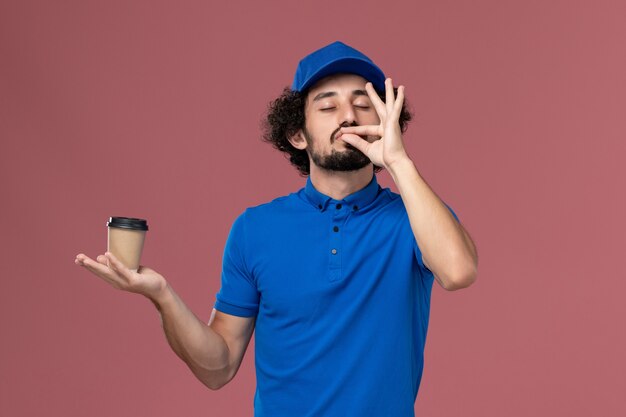 青い制服とピンクの壁に彼の手に配達コーヒーカップとキャップの男性宅配便の正面図