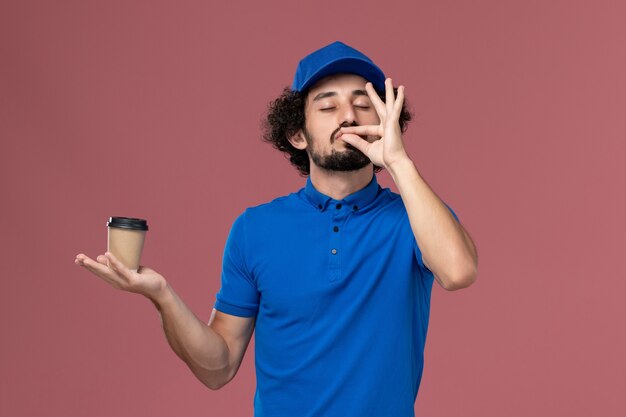 Front view of male courier in blue uniform and cap with delivery coffee cup on his hands on the pink wall