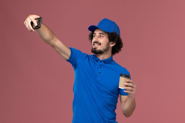 Front view of male courier in blue uniform and cap with delivery coffee cup on his hands taking photo on pink wall