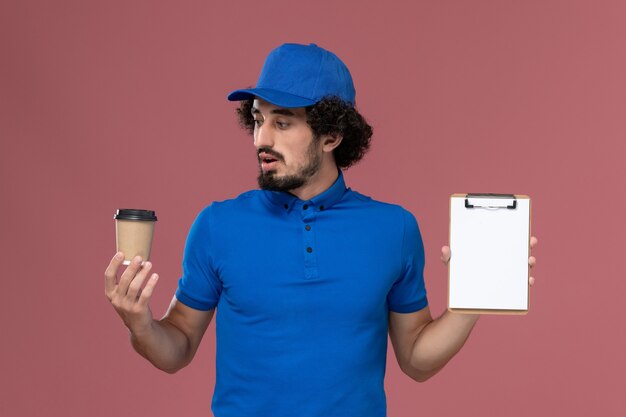 Foto gratuita vista frontale del corriere maschio in uniforme blu e cappuccio con tazza di caffè di consegna e blocco note sulle mani sul muro rosa