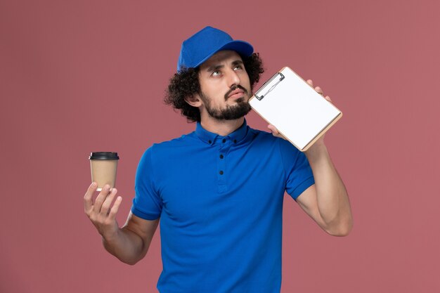 Front view of male courier in blue uniform and cap with delivery coffee cup and notepad on his hands thinking on the pink wall