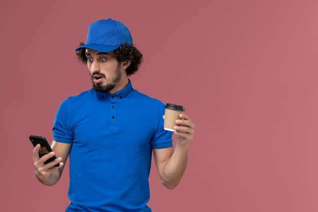 Вид спереди курьера-мужчины в синей форме и кепке с доставкой кофейной чашки и телефона на руках на розовой стене