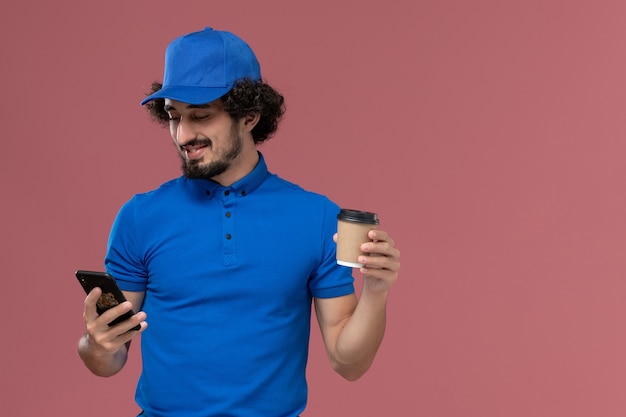 Vista frontale del corriere maschio in uniforme blu e cappuccio con la tazza di caffè di consegna e il telefono sulle mani sul muro rosa