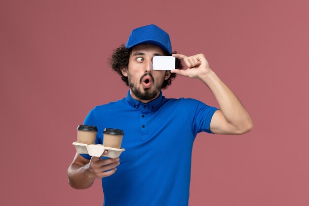 Front view of male courier in blue uniform and cap with delivery coffee cups and card on his hands on the pink wall