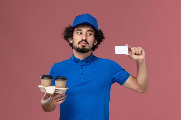 분홍색 벽에 그의 손에 배달 커피 컵과 카드와 파란색 유니폼과 모자 남성 택배의 전면보기
