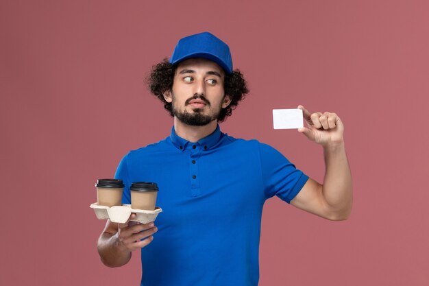 青いユニフォームとピンクの壁に彼の手に配達コーヒーカップとカードとキャップの男性の宅配便の正面図