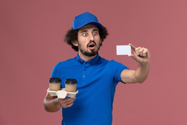 Вид спереди курьера-мужчины в синей форме и кепке с доставкой кофейных чашек и открыткой на руках на розовой стене