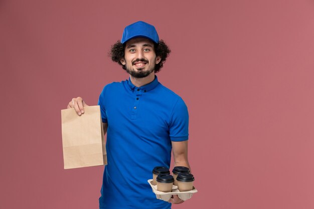 Front view of male courier in blue uniform cap with delivery coffee cups and food package on his hands on pink wall