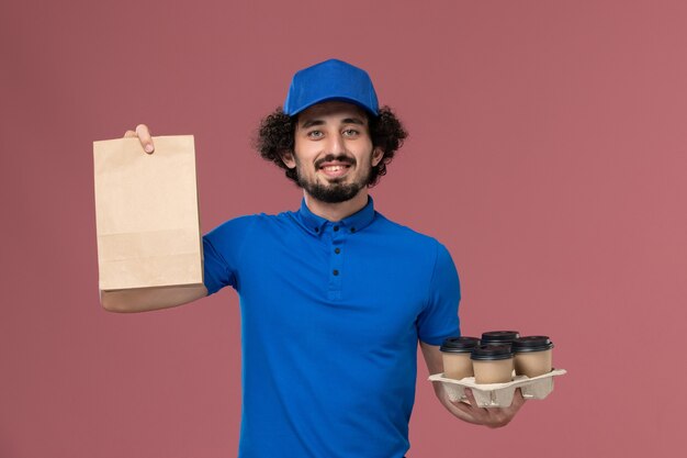 Вид спереди курьера-мужчины в синей форменной кепке с доставкой кофейных чашек и продуктового пакета на руках на розовой стене