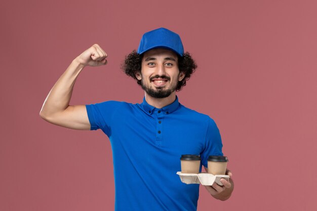 青いユニフォームとピンクの壁で曲がっている彼の手に配達コーヒーカップとキャップの男性の宅配便の正面図