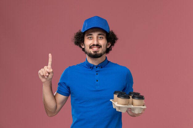 淡いピンクの壁に彼の手に配達コーヒーカップと青い制服キャップの男性宅配便の正面図