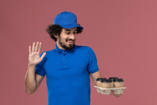 Front view of male courier in blue uniform and cap with delivery coffee cups on his hands on light-pink wall