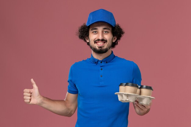 Вид спереди курьера-мужчины в синей форме и кепке с доставочными кофейными чашками на руках на светло-розовой стене