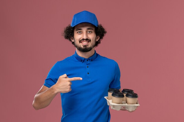 Front view of male courier in blue uniform cap with delivery coffee cups on his hands on light-pink wall