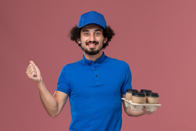 Вид спереди курьера-мужчины в синей форме и кепке с доставочными кофейными чашками на руках на светло-розовой стене