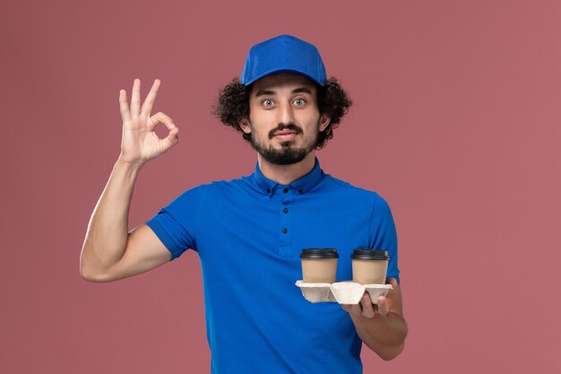 Front view of male courier in blue uniform and cap with delivery coffee cups on his hands on pink wall