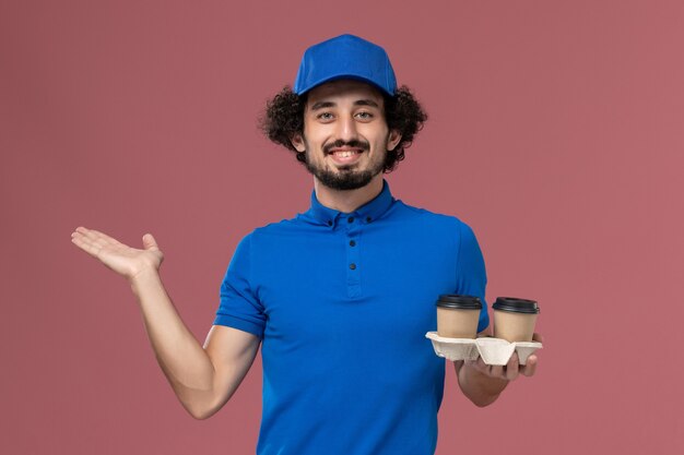 Вид спереди курьера-мужчины в синей форме и кепке с доставленными кофейными чашками на руках на розовой стене
