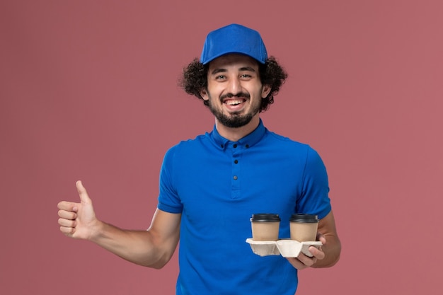 Front view of male courier in blue uniform and cap with delivery coffee cups on his hands on pink wall