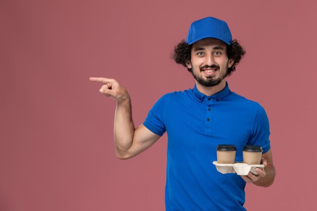 Vista frontale del corriere maschio in protezione uniforme blu con tazze di caffè di consegna sulle sue mani sulla parete rosa