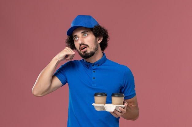 Вид спереди курьера-мужчины в синей форме и кепке с доставленными кофейными чашками на руках на розовой стене