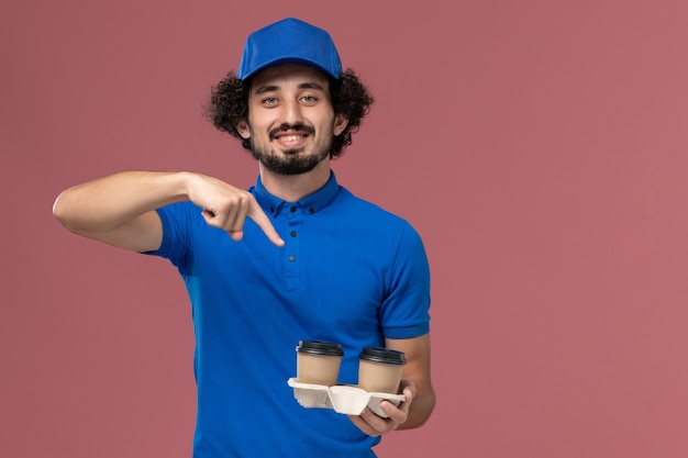 Front view of male courier in blue uniform and cap with delivery coffee cups on his hands on pink wall
