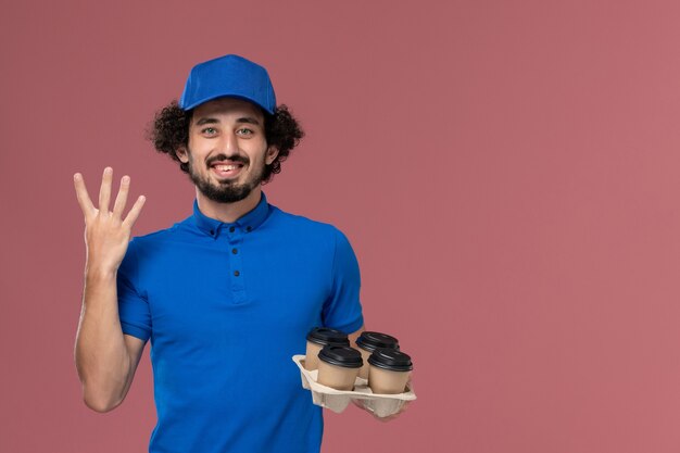 Вид спереди курьера-мужчины в синей униформе с доставкой кофейных чашек на руках, улыбаясь на розовой стене