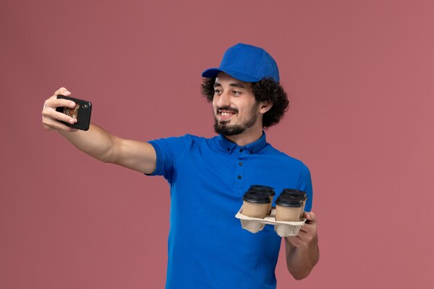 Front view of male courier in blue uniform cap with delivery coffee cups on his hands taking picture on pink wall