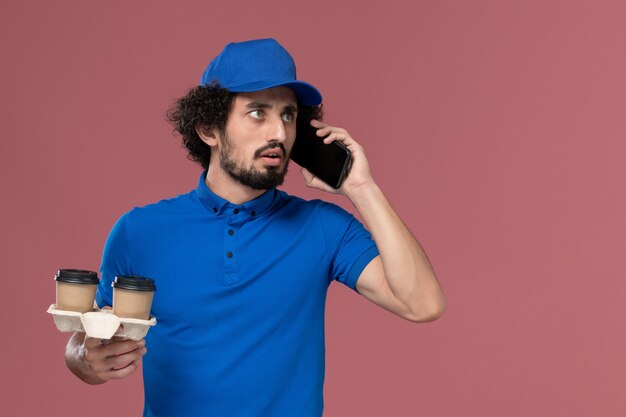 Front view of male courier in blue uniform and cap with delivery coffee cups on his hands talking on the phone on pink wall