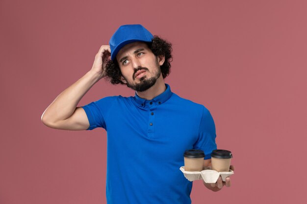 Front view of male courier in blue uniform and cap with delivery coffee cups on his hands thinking on pink wall