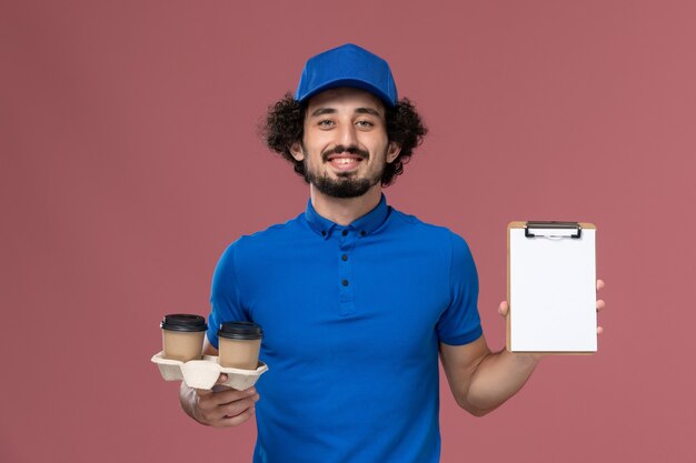 青いユニフォームとピンクの壁に彼の手に配達コーヒーカップとメモ帳とキャップの男性宅配便の正面図