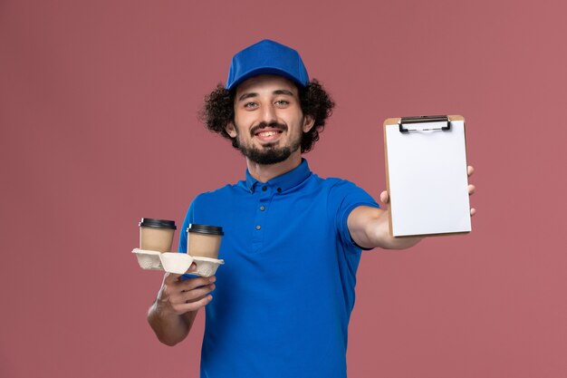 青いユニフォームとピンクの壁に彼の手に配達コーヒーカップとメモ帳とキャップの男性宅配便の正面図