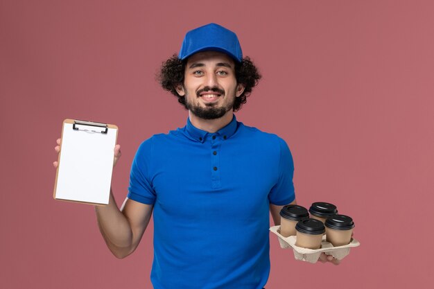Front view of male courier in blue uniform cap with delivery coffee cups and notepad on his hands on pink wall