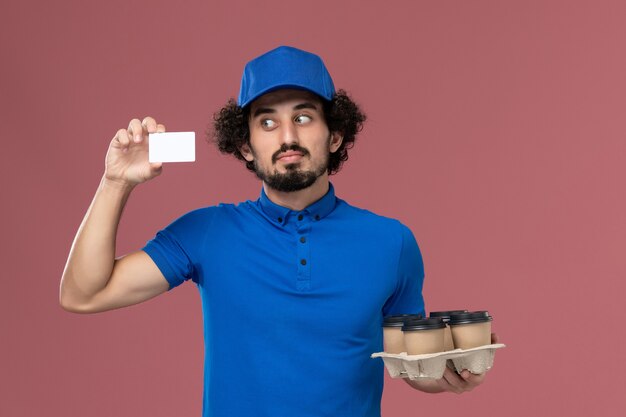 Front view of male courier in blue uniform cap with delivery coffee cups and plastic card on his hands on pink wall