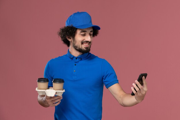 青い制服を着た男性の宅配便の正面図とピンクの壁に彼の手に配達コーヒーカップと仕事の電話とキャップ