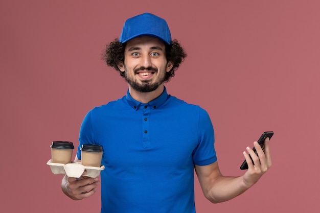 Front view of male courier in blue uniform and cap with delivery coffee cups and work phone on his hands on the pink wall