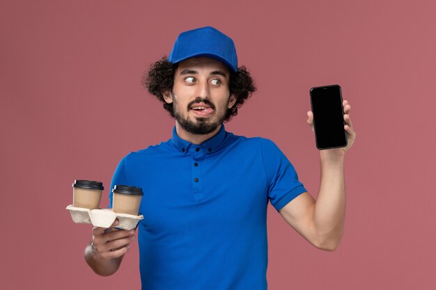 Front view of male courier in blue uniform and cap with delivery coffee cups and work phone on his hands