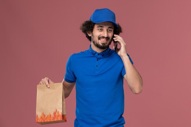 Front view of male courier in blue uniform cap with delivery food package on his hands talking on the phone on pink wall