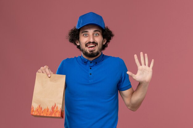 淡いピンクの壁に彼の手に配達紙食品パッケージと青い制服キャップの男性宅配便の正面図