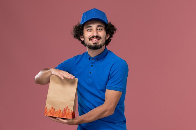 Front view of male courier in blue uniform cap with delivery paper food package on his hands on the light pink wall