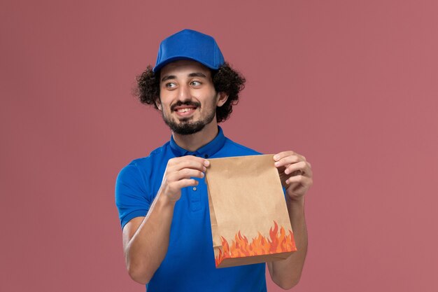 Front view of male courier in blue uniform cap with delivery paper food package on his hands on the light pink wall