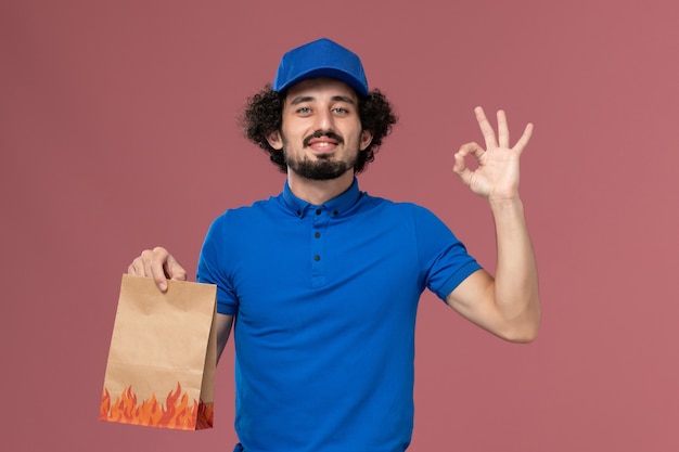 Front view of male courier in blue uniform cap with delivery paper food package on his hands on the light pink wall