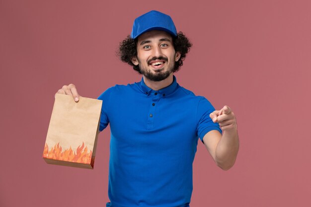 Front view of male courier in blue uniform cap with delivery paper food package on his hands smiling on the light pink wall
